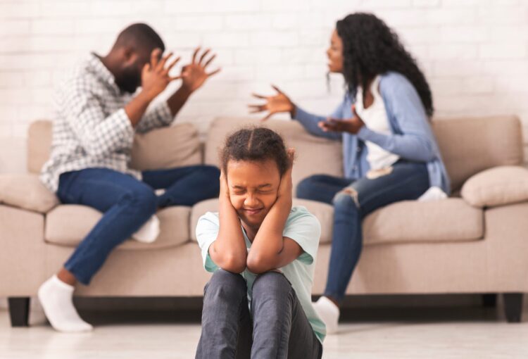 Little black girl closing ears with hands not to listen parents quarrel, sitting on floor alone, selective focus on child