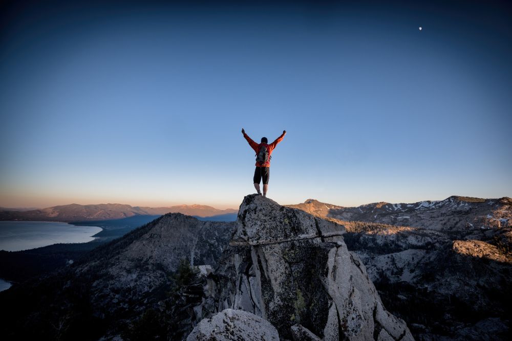 Person standing triumphantly on a mountaintop at sunrise, arms raised in victory, overlooking a vast landscape of mountains and water.
