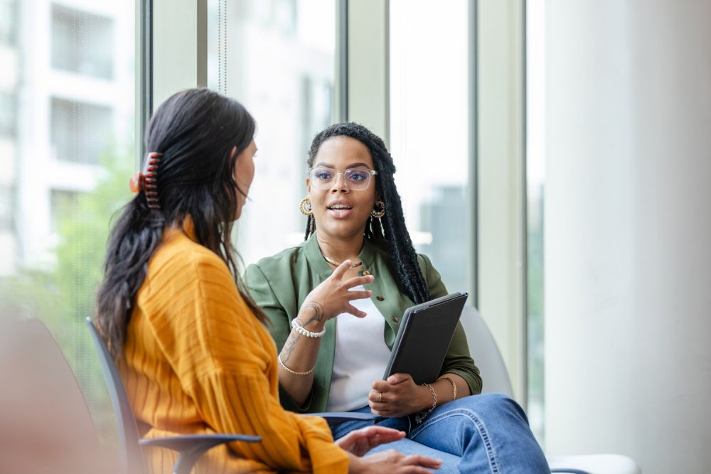 Two women sitting by large windows having a supportive conversation, with one holding a tablet and gesturing while the other listens attentively.
