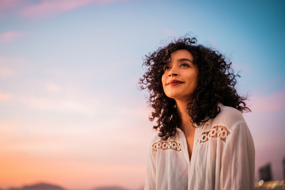 Woman standing outdoors at sunset with a soft smile and peaceful expression, symbolizing gratitude, hope, and emotional well-being.