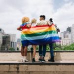 Four people stand arm in arm on a city overlook, draped in a large rainbow Pride flag. Tall buildings and greenery are visible in the background.