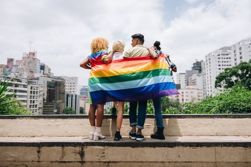 Four people stand arm in arm on a city overlook, draped in a large rainbow Pride flag. Tall buildings and greenery are visible in the background.