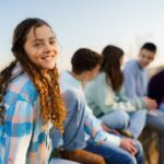 A smiling teenage girl in a plaid shirt sits outdoors on a stone wall with a group of other teens, enjoying time together on a sunny day.