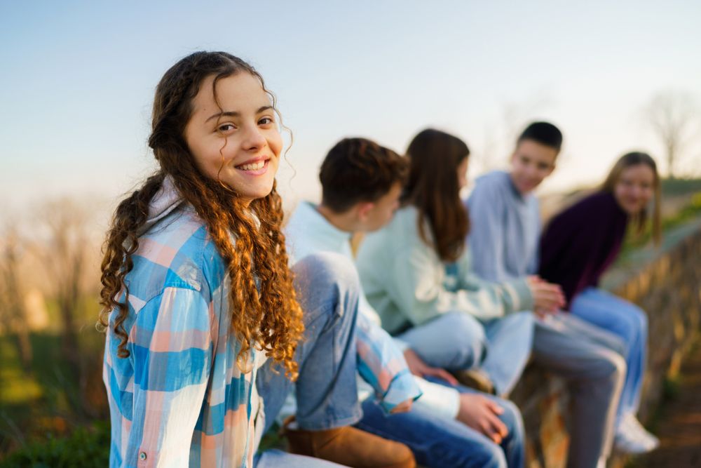 A smiling teenage girl in a plaid shirt sits outdoors on a stone wall with a group of other teens, enjoying time together on a sunny day.
