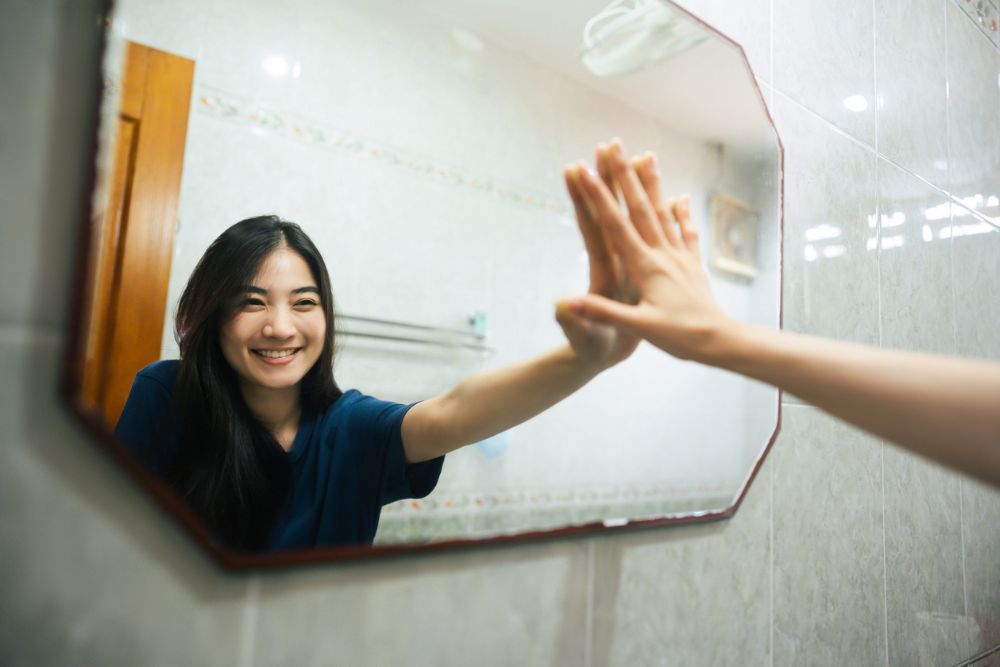 Young adult smile woman practice self talk conversation in the morning in bathroom at home. Hand hi-touch at mirror. Healthy lifestyle after wake up concept.