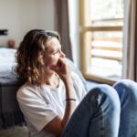 A woman sits on the floor by her bed with her knees pulled up, resting her hand on her chin. She looks out the window thoughtfully, appearing worried or reflective.