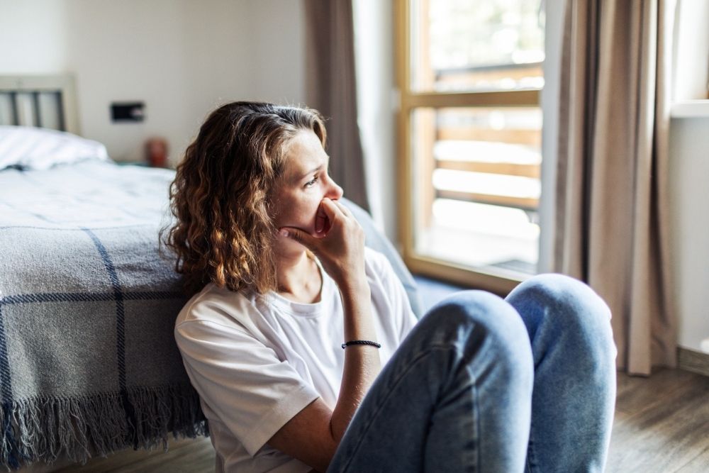 A woman sits on the floor by her bed with her knees pulled up, resting her hand on her chin. She looks out the window thoughtfully, appearing worried or reflective.