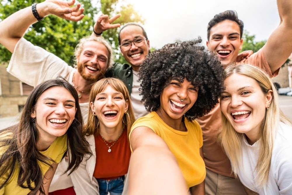 Group of diverse young adults smiling and posing closely together for a selfie outdoors, expressing joy, connection, and positive summer energy.