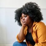 Woman sitting indoors with her hand on her face, looking thoughtful and sad, representing symptoms of depression.
