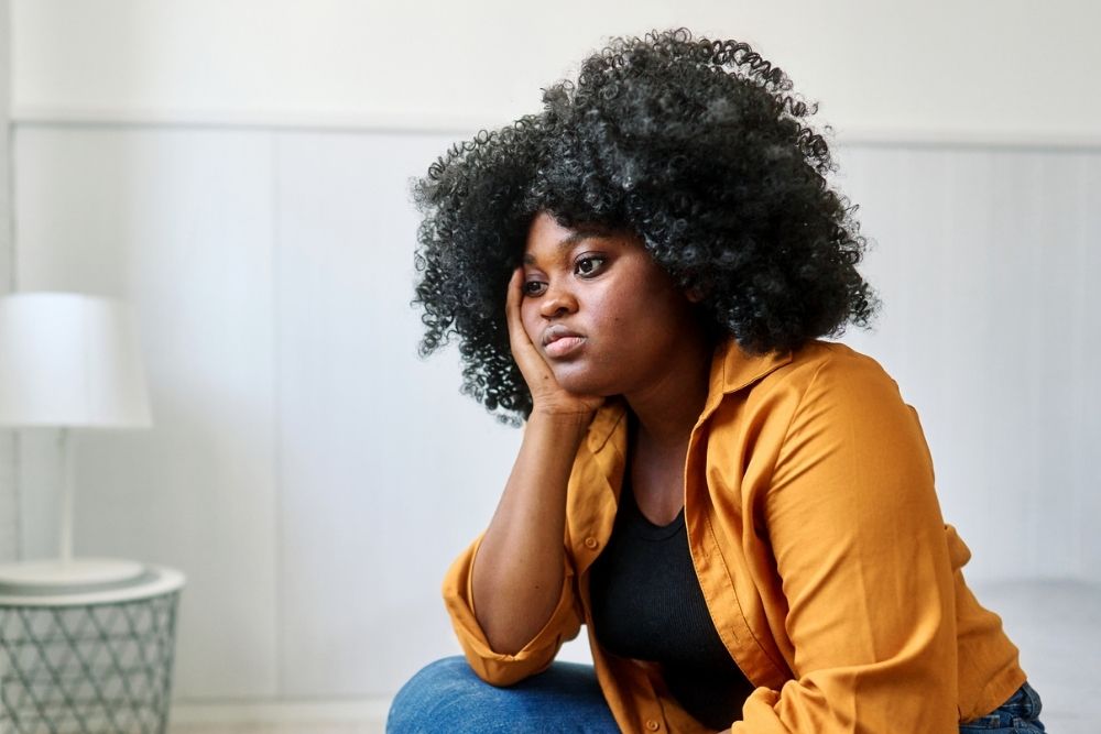 Woman sitting indoors with her hand on her face, looking thoughtful and sad, representing symptoms of depression.