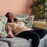 Woman reclining on a couch with her head tilted back and eyes closed, appearing exhausted or unmotivated in a home setting.