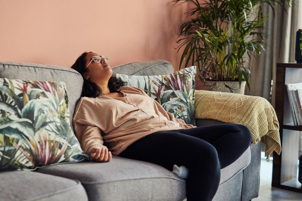 Woman reclining on a couch with her head tilted back and eyes closed, appearing exhausted or unmotivated in a home setting.