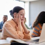 A student sits in a classroom resting her face in her hands, looking tired or distracted while working on a laptop.