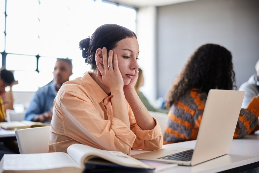 A student sits in a classroom resting her face in her hands, looking tired or distracted while working on a laptop.