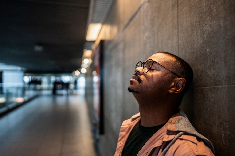 Man leaning against a wall with eyes closed, appearing stressed or deep in thought in a dimly lit hallway.