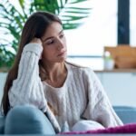 A young woman sits on a couch with a thoughtful, slightly sad expression, resting her head on her hand in a bright home setting.