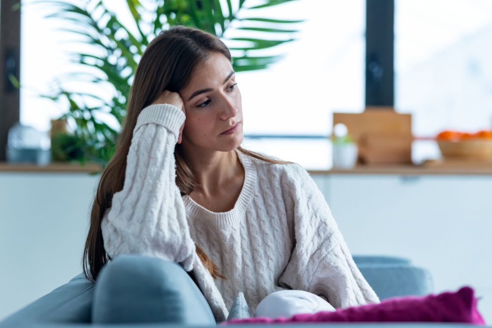 A young woman sits on a couch with a thoughtful, slightly sad expression, resting her head on her hand in a bright home setting.