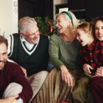 A family sits together indoors during the holidays, laughing and smiling. An older couple, a young girl, and two adults enjoy a warm, joyful moment.
