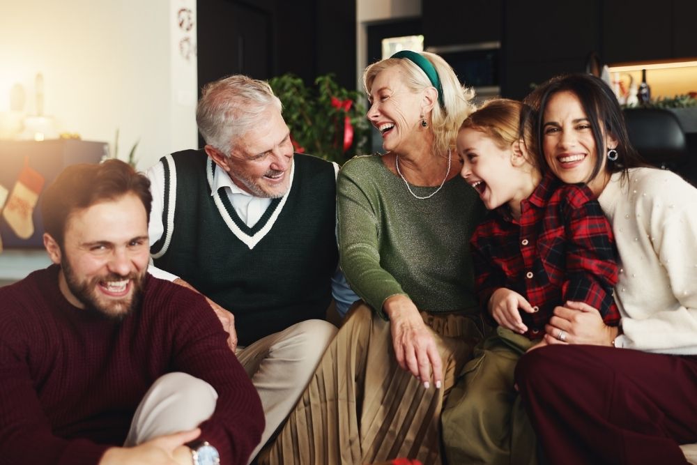 A family sits together indoors during the holidays, laughing and smiling. An older couple, a young girl, and two adults enjoy a warm, joyful moment.