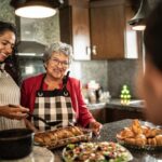 Two women wearing aprons preparing holiday food in a kitchen, smiling while serving a baked dish, with festive decorations in the background.