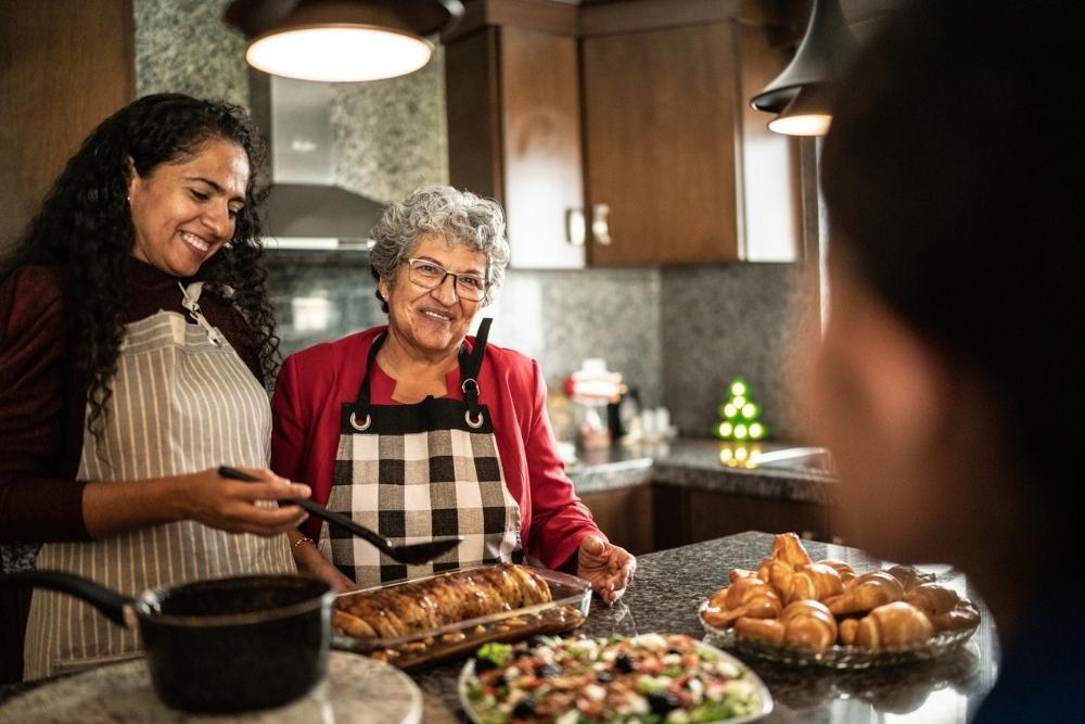 Two women wearing aprons preparing holiday food in a kitchen, smiling while serving a baked dish, with festive decorations in the background.