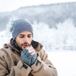 A man bundled in winter clothing stands outdoors in a snowy landscape, breathing into his hands to stay warm on a cold day.