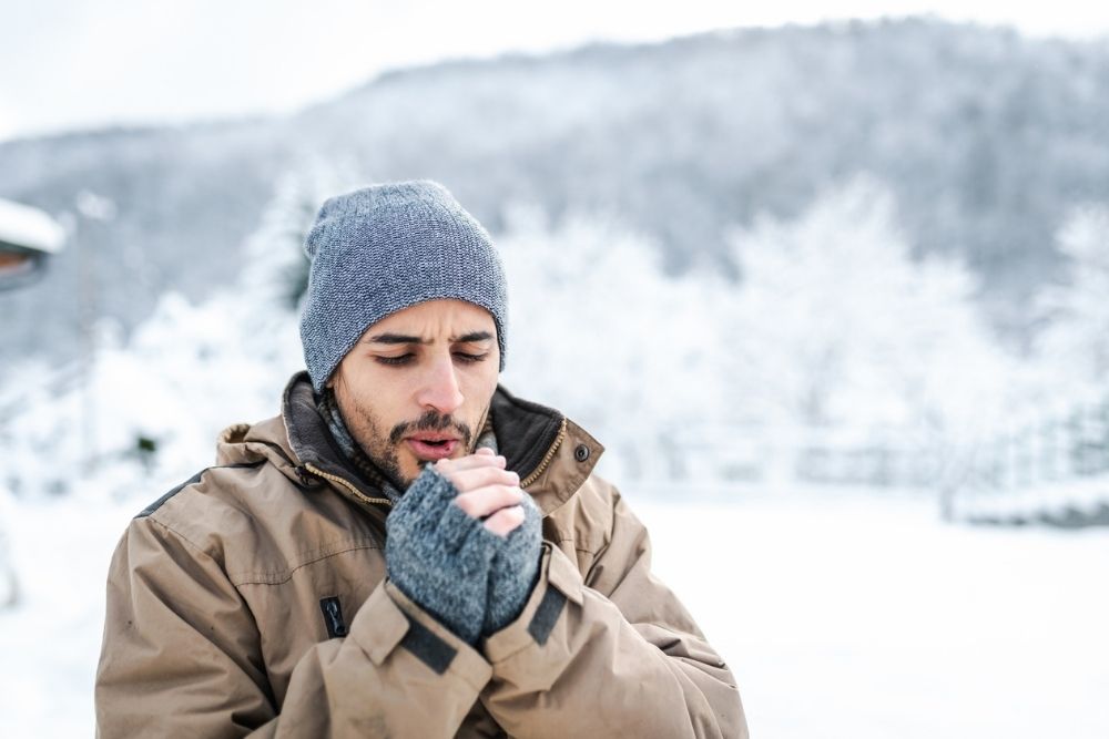A man bundled in winter clothing stands outdoors in a snowy landscape, breathing into his hands to stay warm on a cold day.