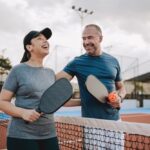 Two adults smiling and talking while holding pickleball paddles on an outdoor court, enjoying light physical activity together.