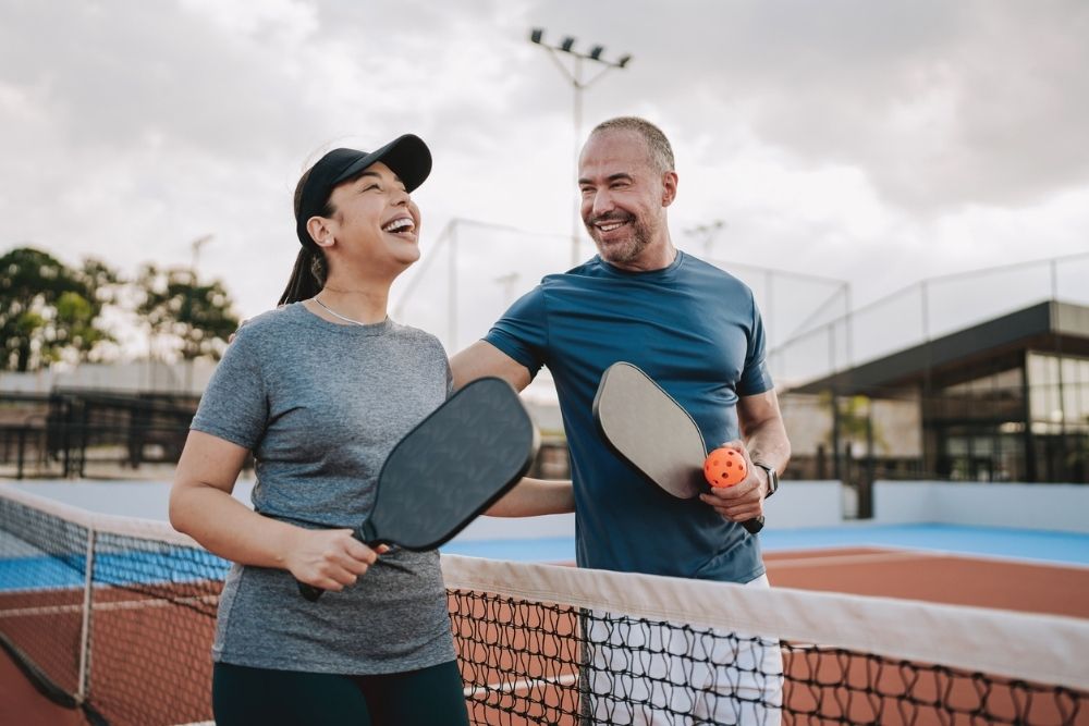 Two adults smiling and talking while holding pickleball paddles on an outdoor court, enjoying light physical activity together.