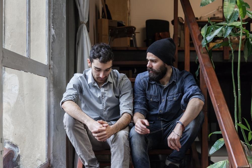 Two men sit on wooden steps indoors. The man on the left looks down, appearing contemplative, while the man on the right speaks to him supportively knowing what to say to someone who is struggling with depression.